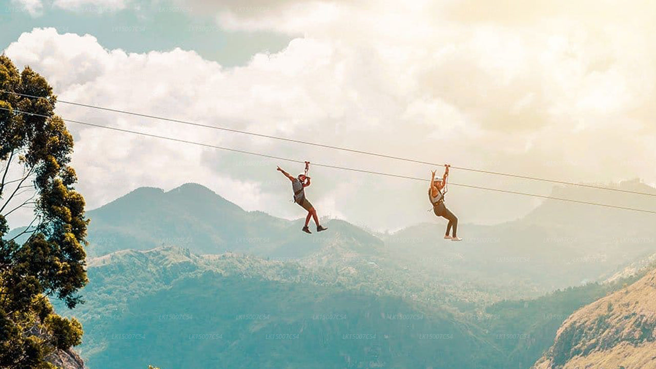 Two people ziplining across lush green mountains with a backdrop of blue sky and clouds.