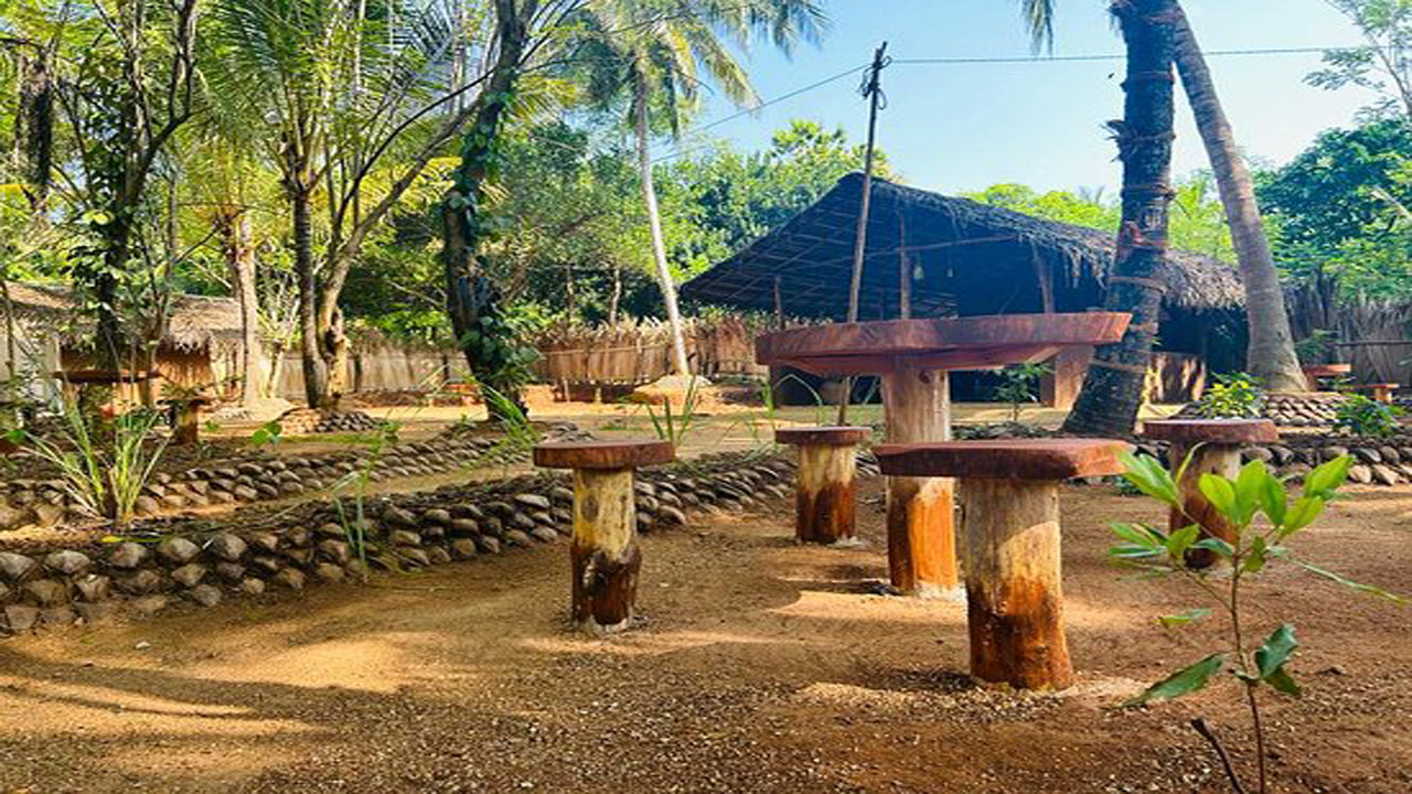 A group of people standing around a village center with wooden structures and a thatched roof structure, surrounded by lush greenery.